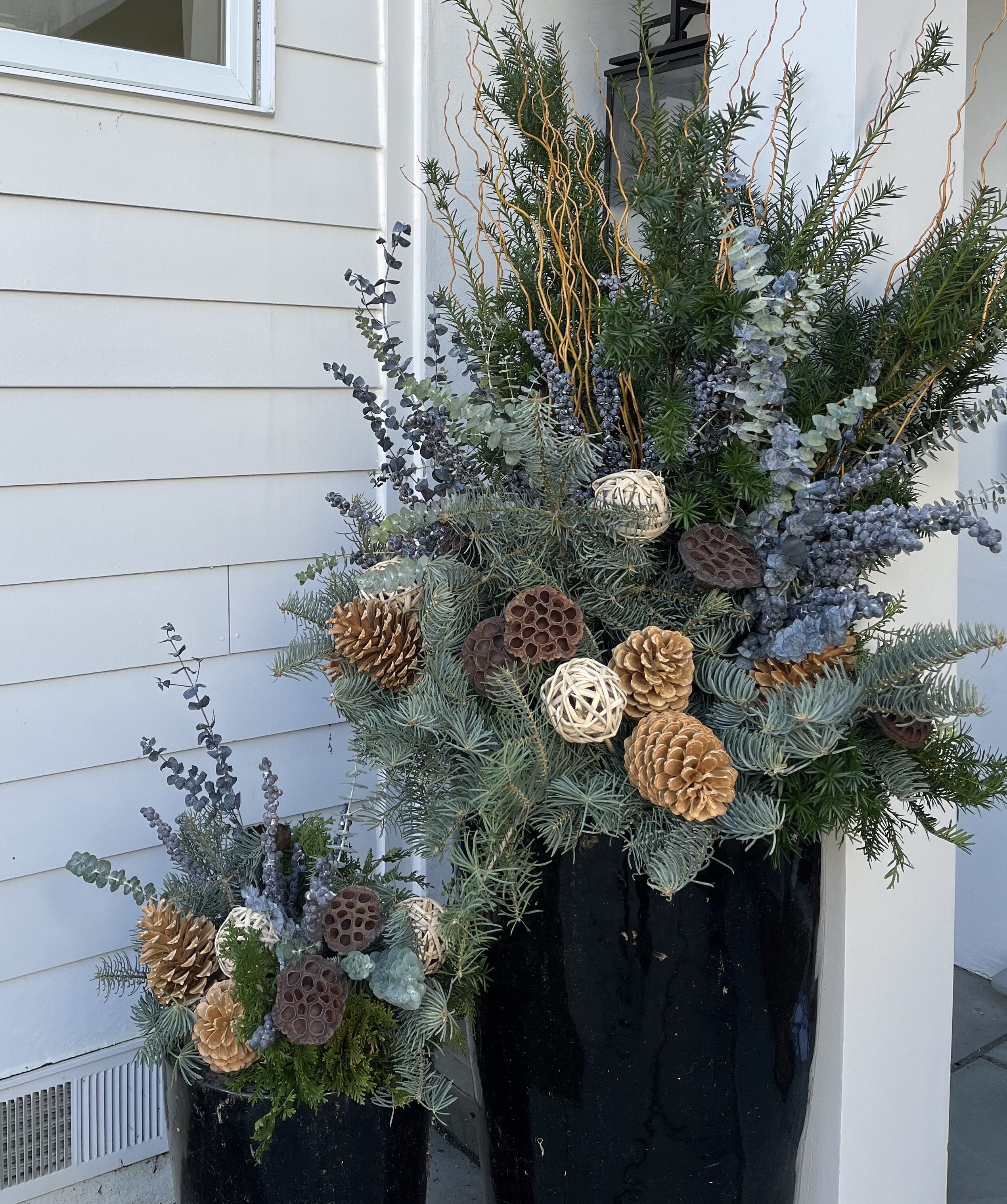 2 matching black planters with pinecones, eucalyptus, and other winter greenery alongside the exterior of a white building.