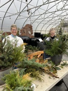 2 people in sweaters building large winter planters inside a greenhouse.