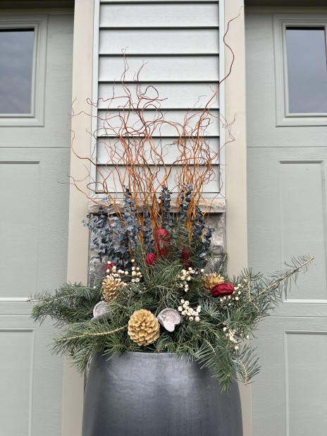 Large metallic silver planter with an assortment of green, brown, red, and white winter greenery sits in front of a house.