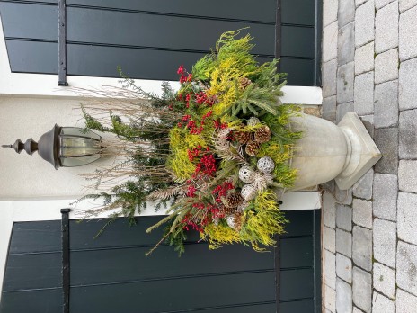 Large stone planter full of winter greenery, pine cones, and berries sits in a stone driveway.